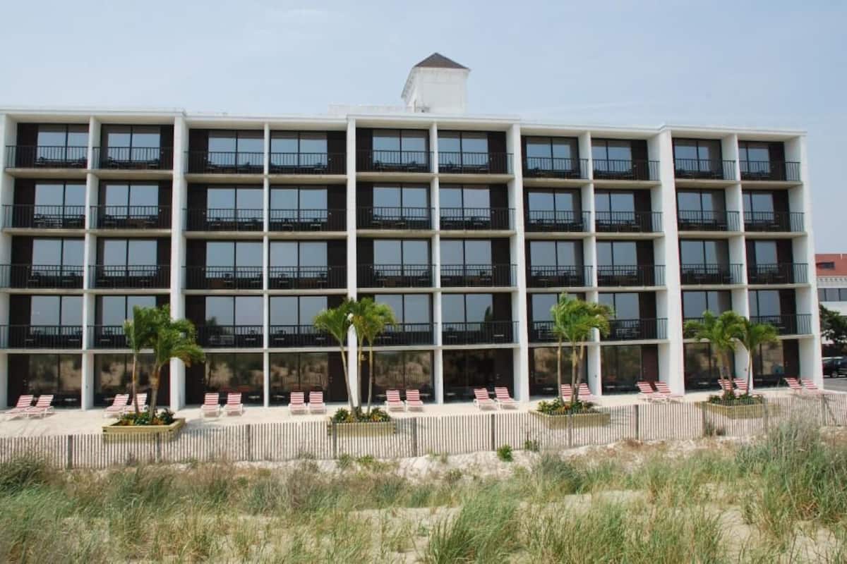 A multi-story hotel with balconies, a sandy beach area with lounge chairs, and palm trees.