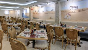 A restaurant with multiple dining tables and wicker chairs, featuring a beach mural on the wall.