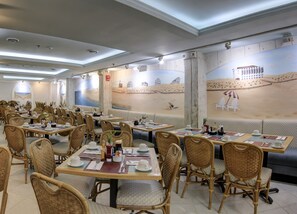 A restaurant with multiple dining tables and wicker chairs, featuring a beach mural on the wall.