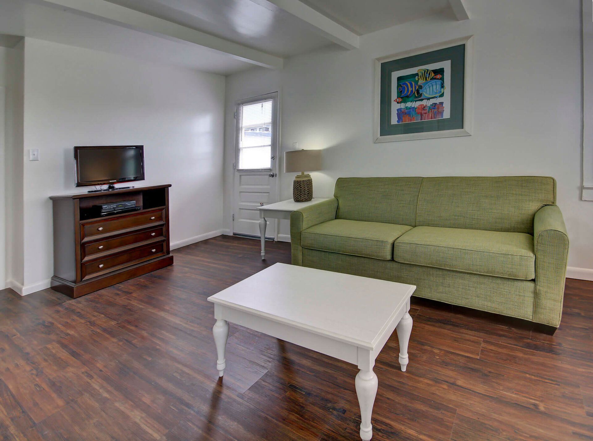A living room with a green sofa, a white coffee table, a TV on a wooden stand, and a painting on the wall.