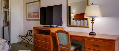 Habitación de hotel con una cama, un escritorio con televisor, una silla, un aparador y una lámpara.