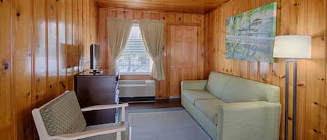 Una habitación con paredes de madera, un sofá, una silla, un escritorio con televisor y una ventana con cortinas.