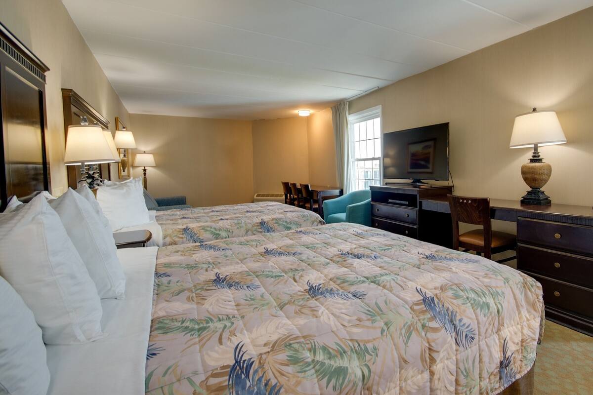 A hotel room with a large bed, two bedside tables with lamps, a desk with a chair, a television, and a window with curtains.