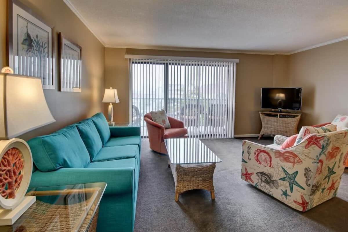 A living room with a teal sofa, a red armchair, a glass coffee table, a wicker side table, and a television on a stand.