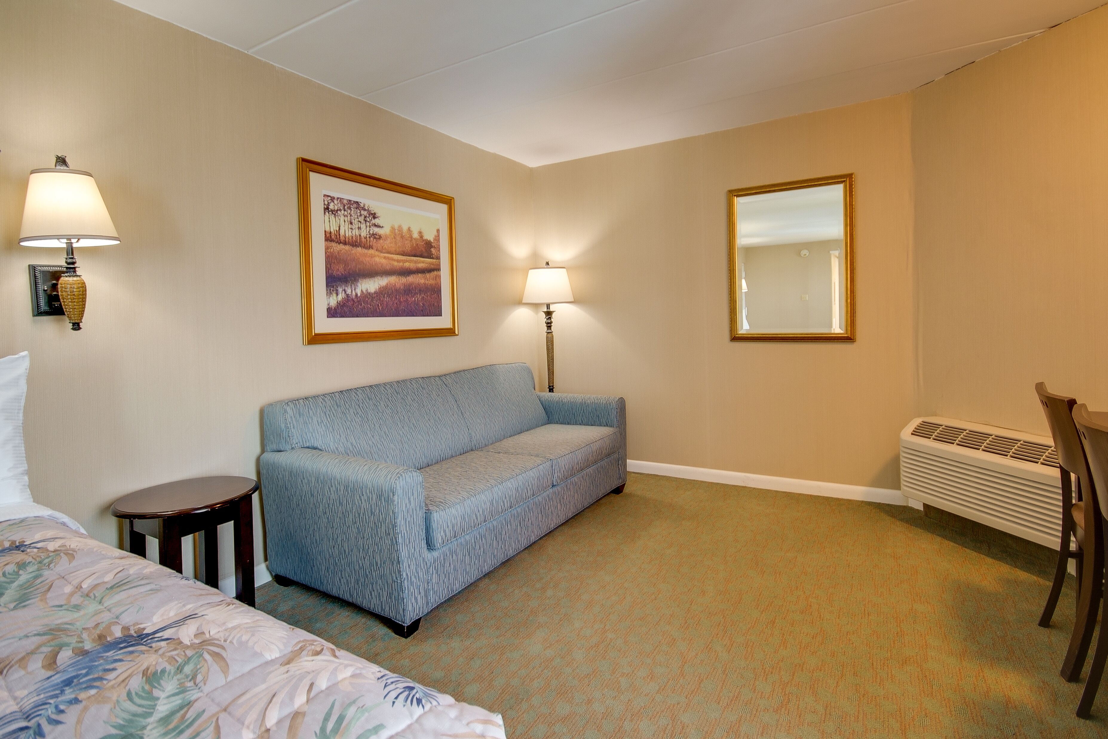 A hotel room with a bed, a blue sofa, two wall-mounted lamps, a framed picture, a radiator, and a chair.