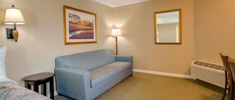 A hotel room with a bed, a blue sofa, two wall-mounted lamps, a framed picture, a radiator, and a chair.