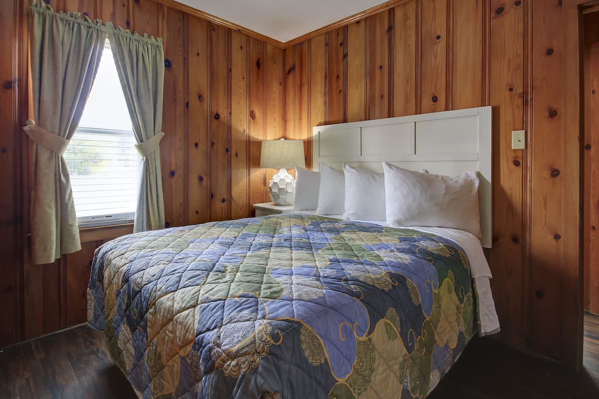 A bedroom with a wooden paneled wall, a bed with a quilted bedspread, a nightstand with a lamp, and a window with curtains.