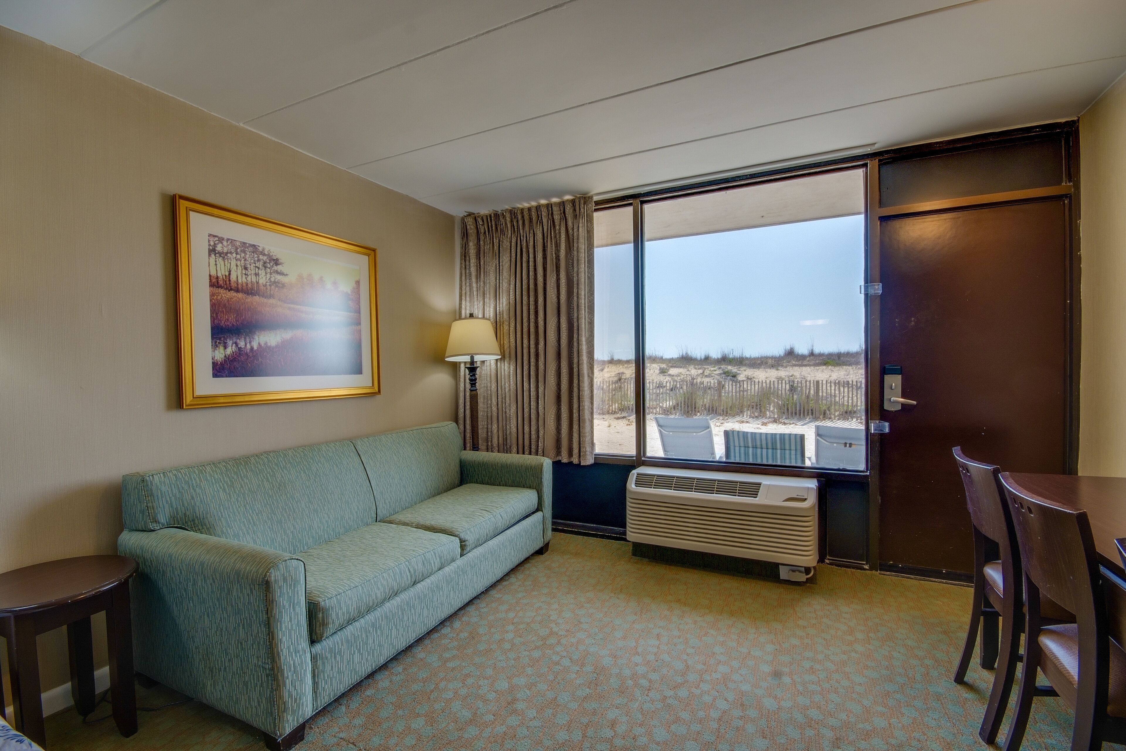 A hotel room with a sofa, a table, a lamp, a large window with a view, and an air conditioning unit.