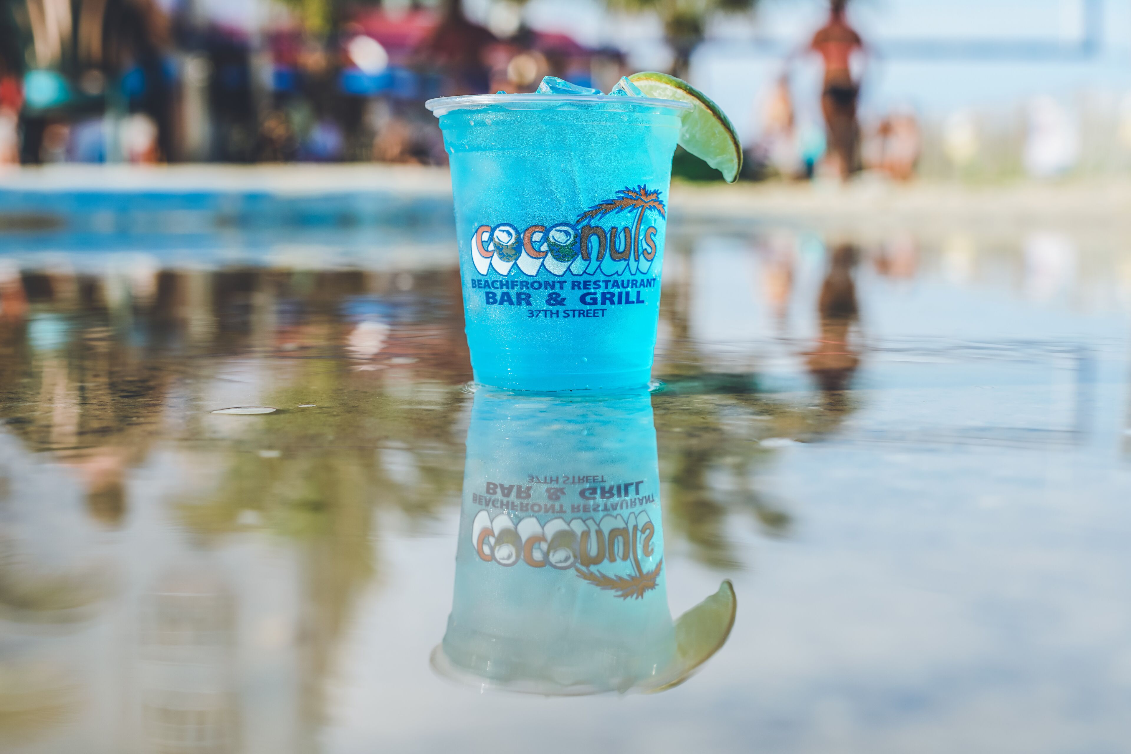 A blue drink with a lime wedge in a plastic cup with the text "coconuts" and "Beachfront Restaurant Bar & Grill 37th Street".