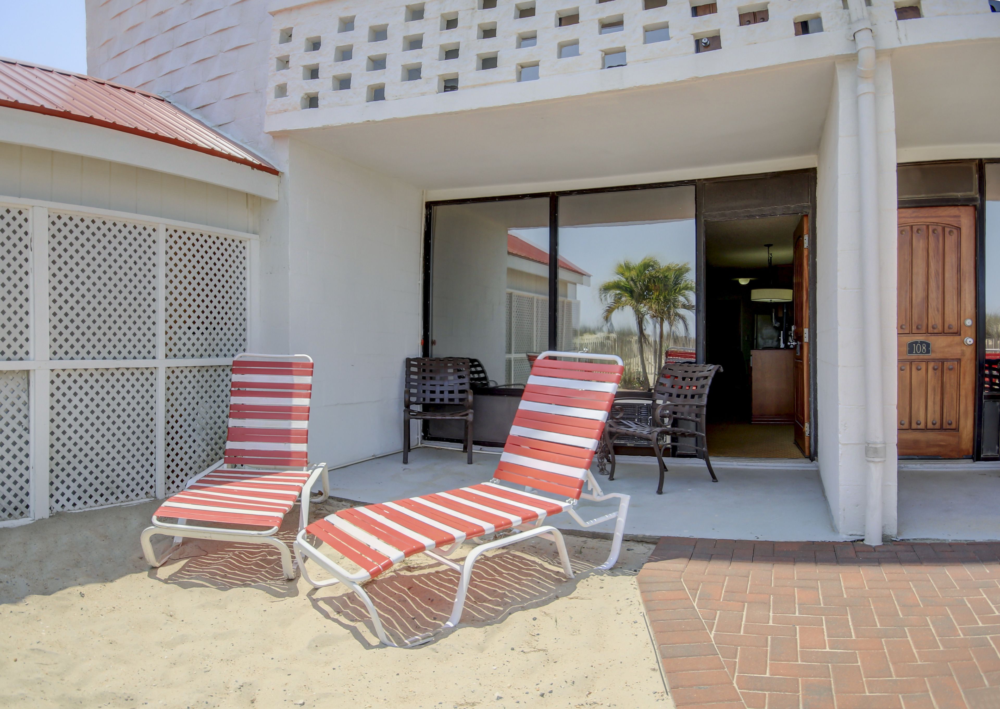 A covered outdoor seating area with two red-striped lounge chairs and a table, adjacent to a building with a white facade and a wooden door marked "108".