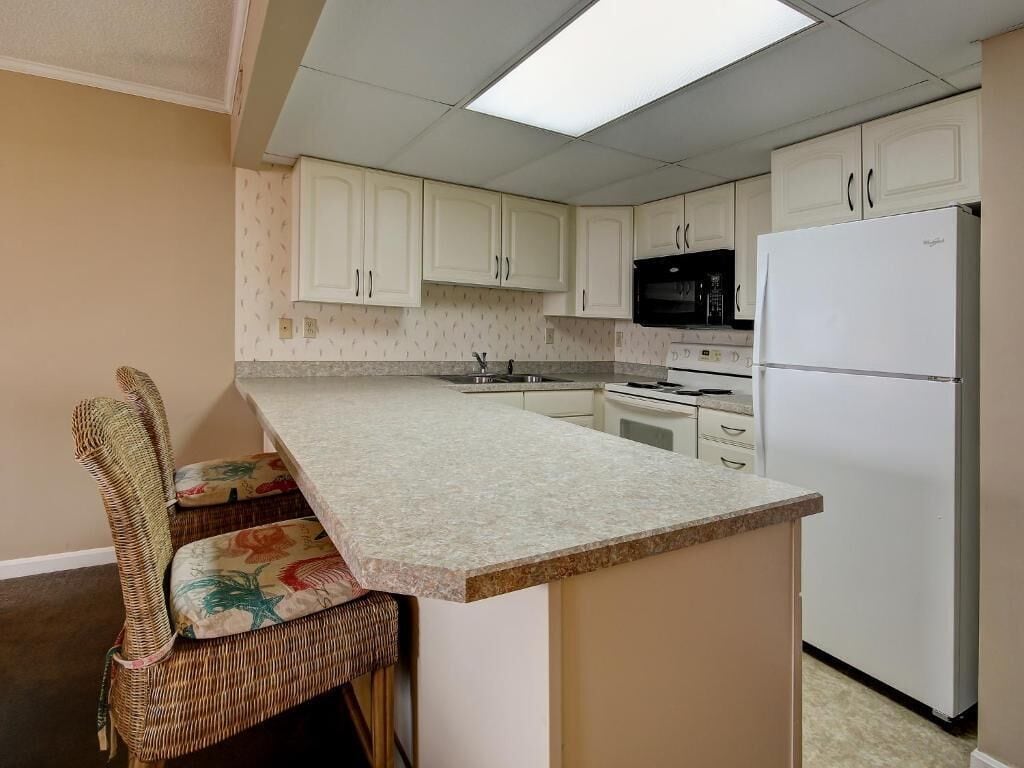 A kitchen with white cabinets, a microwave, a refrigerator, and a dining area with a wicker chair.