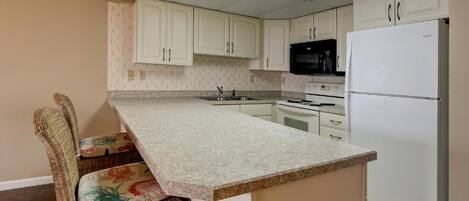 Una cocina con armarios blancos, microondas, refrigerador y una zona de comedor con una silla de mimbre.