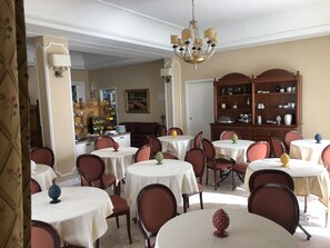 Een eetgedeelte met ronde tafels, stoelen en een kroonluchter. Er is een houten kast met servies en een schilderij aan de muur.