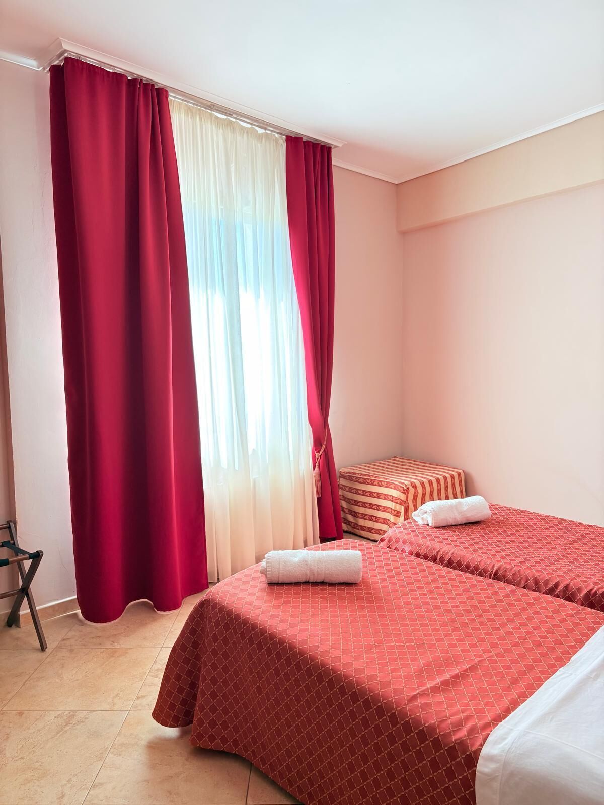A hotel room with two beds, red bedding, a white towel, and a window with red curtains.