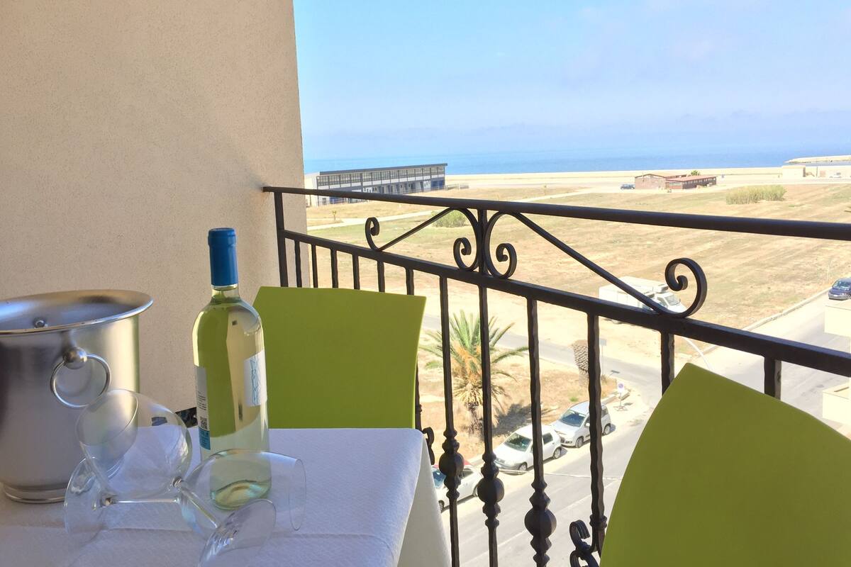 Een balkon met een tafel gedekt voor twee personen, een fles wijn en een pot. De uitzicht is op een strand en een gebouw in de verte.