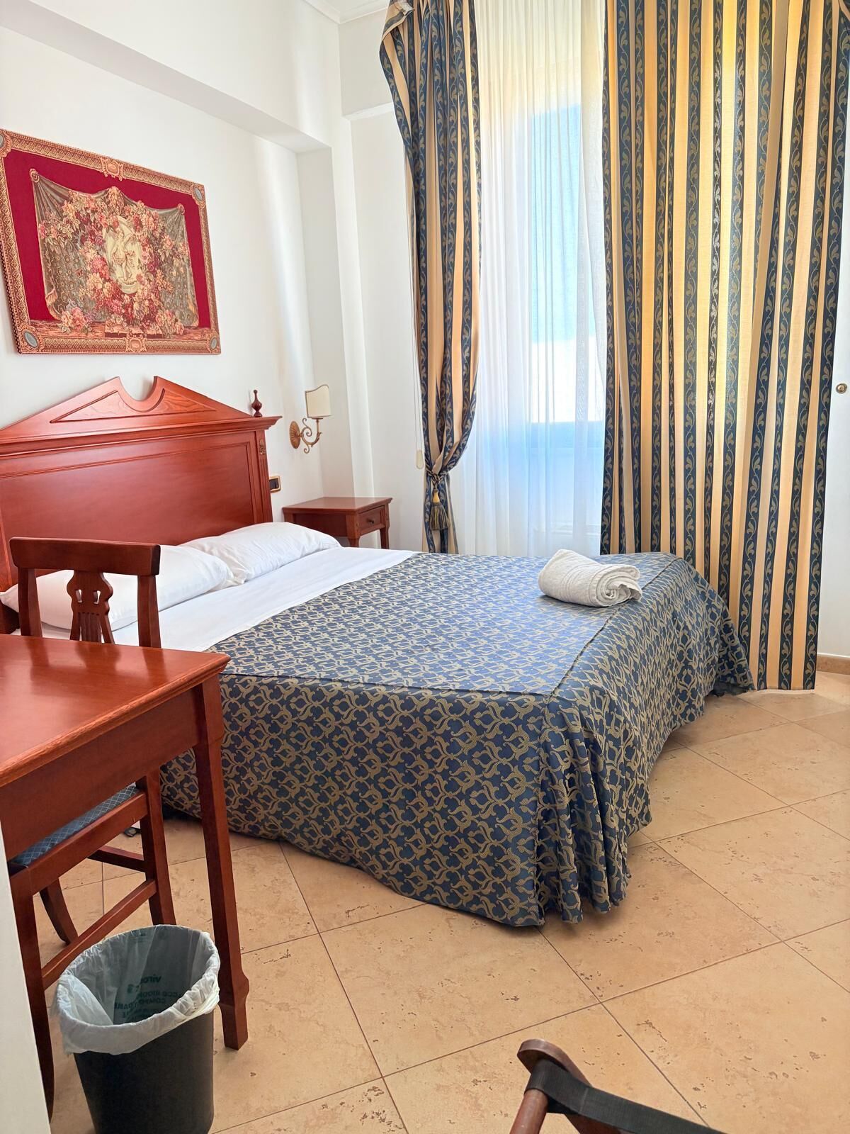 A hotel room with a wooden bed, a nightstand, a desk, and a trash bin.