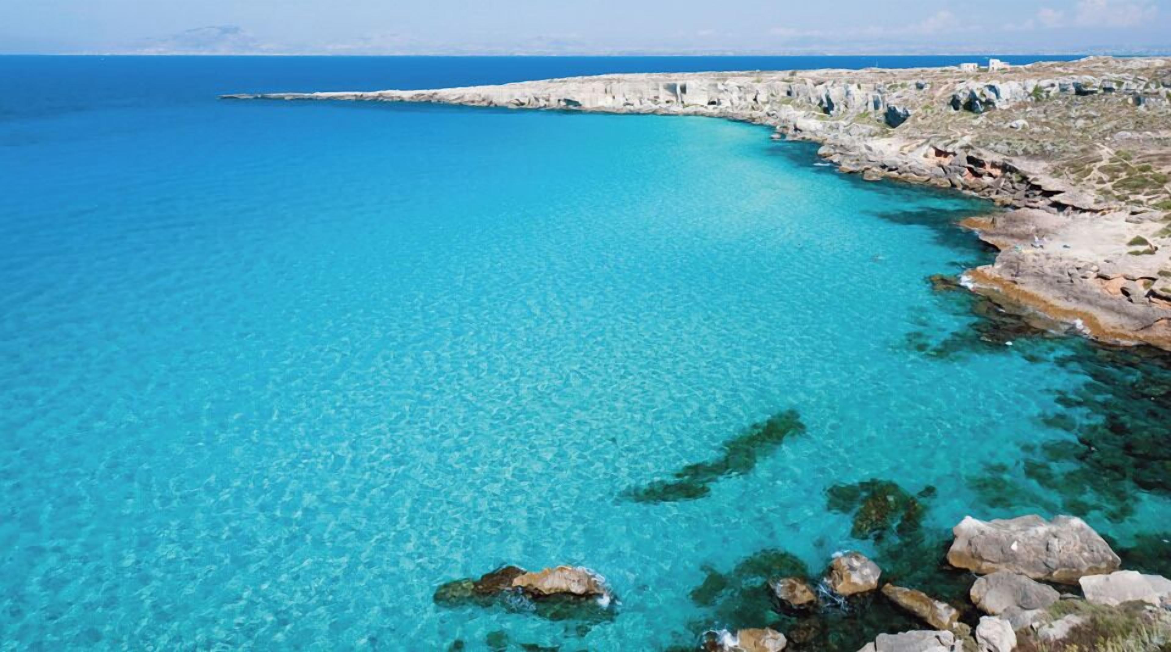 A coastal landscape with clear turquoise waters, rocky shoreline, and a distant view of the sea.