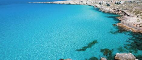 A coastal landscape with clear turquoise waters, rocky shoreline, and a distant view of the sea.