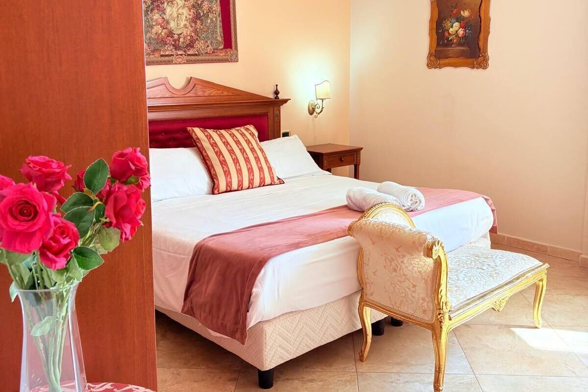 Hotelkamer met een groot bed, een kleiner bed, een stoel, een nachtkastje, een wandlamp en een vaas met bloemen.