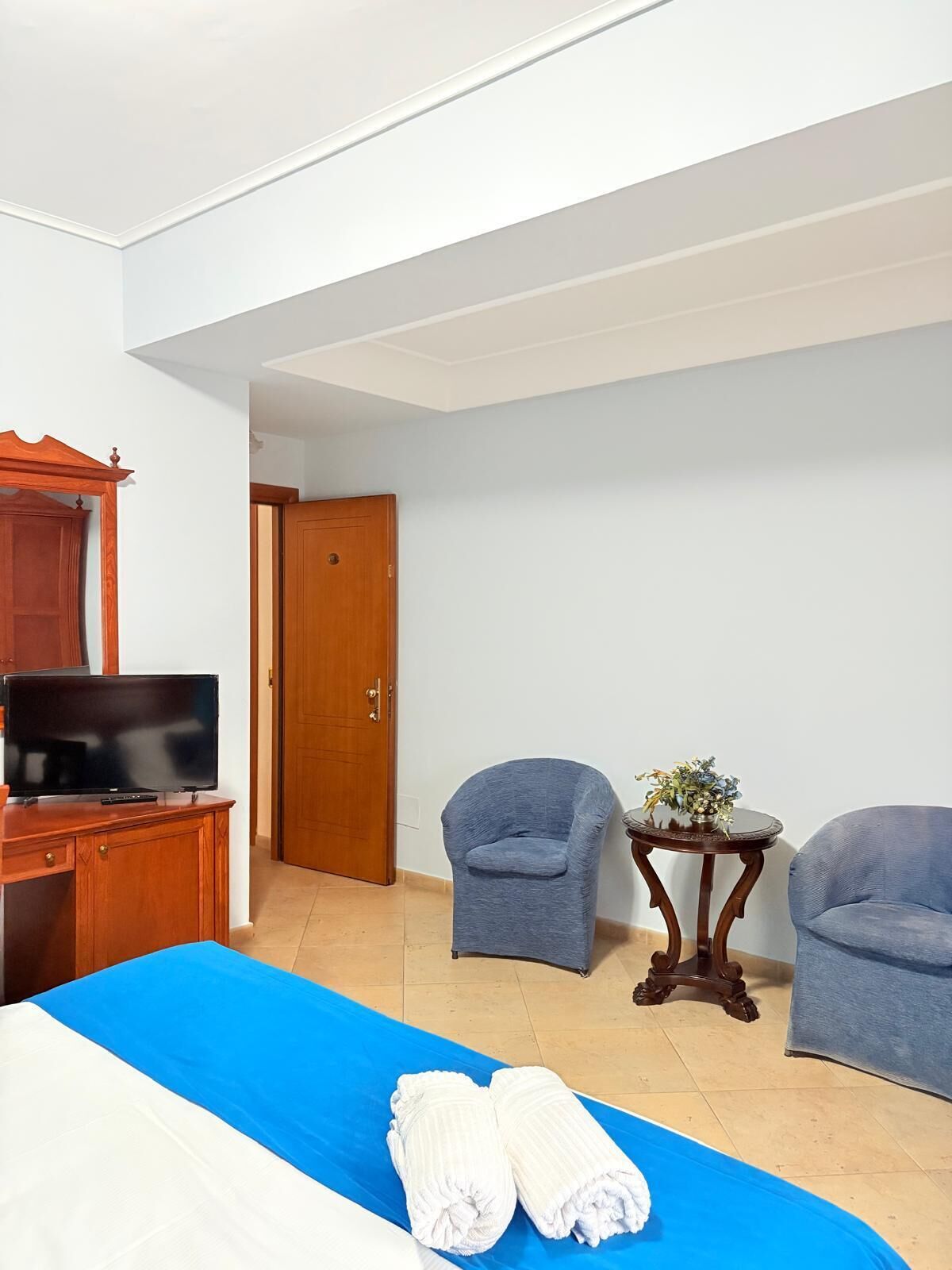 A hotel room with a bed, a TV, two armchairs, a small table with a plant, and a wooden wardrobe.