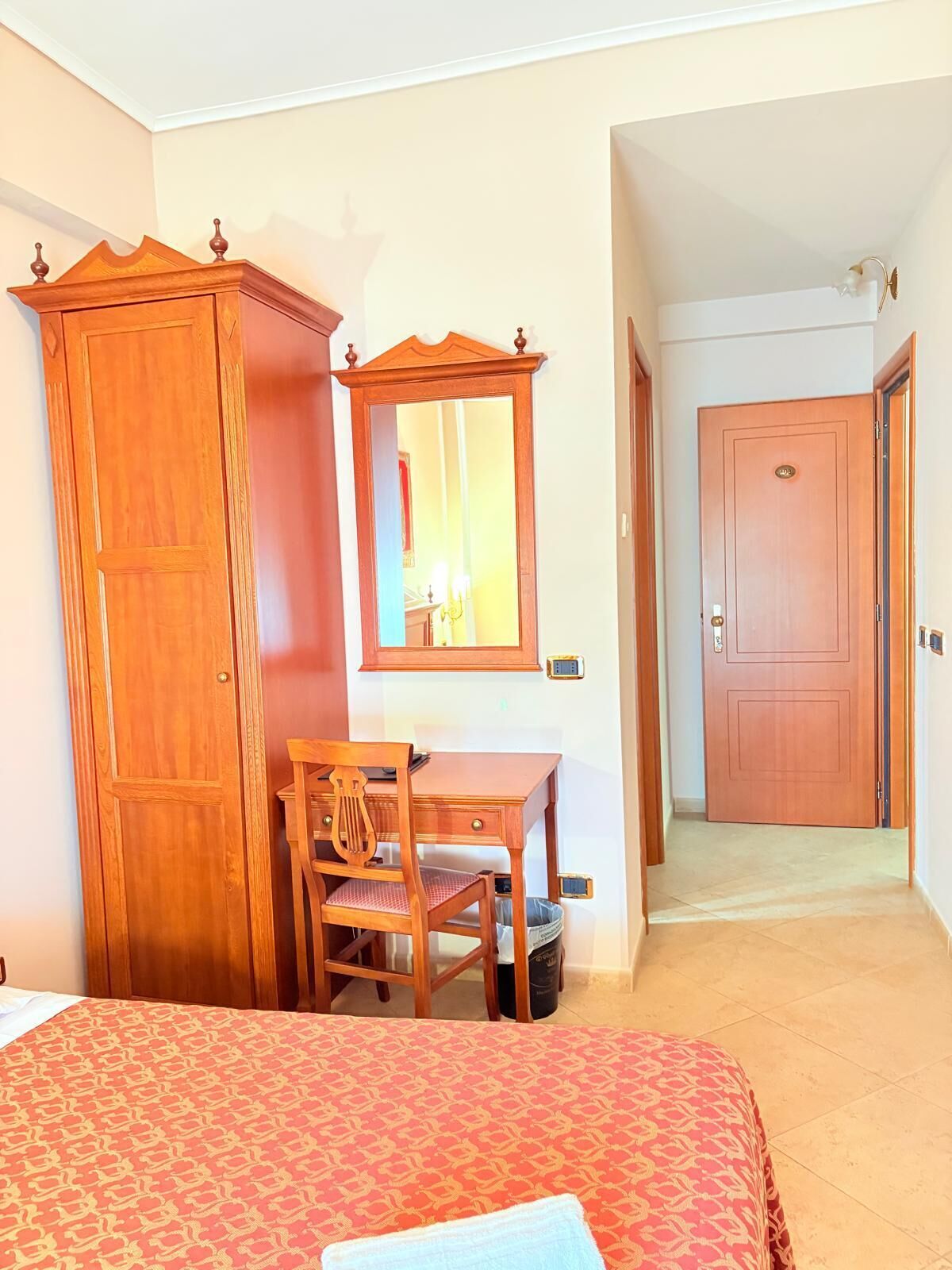 Quarto de hotel com cama, guarda-roupa de madeira, escrivaninha com cadeira, espelho e porta para outro cômodo.