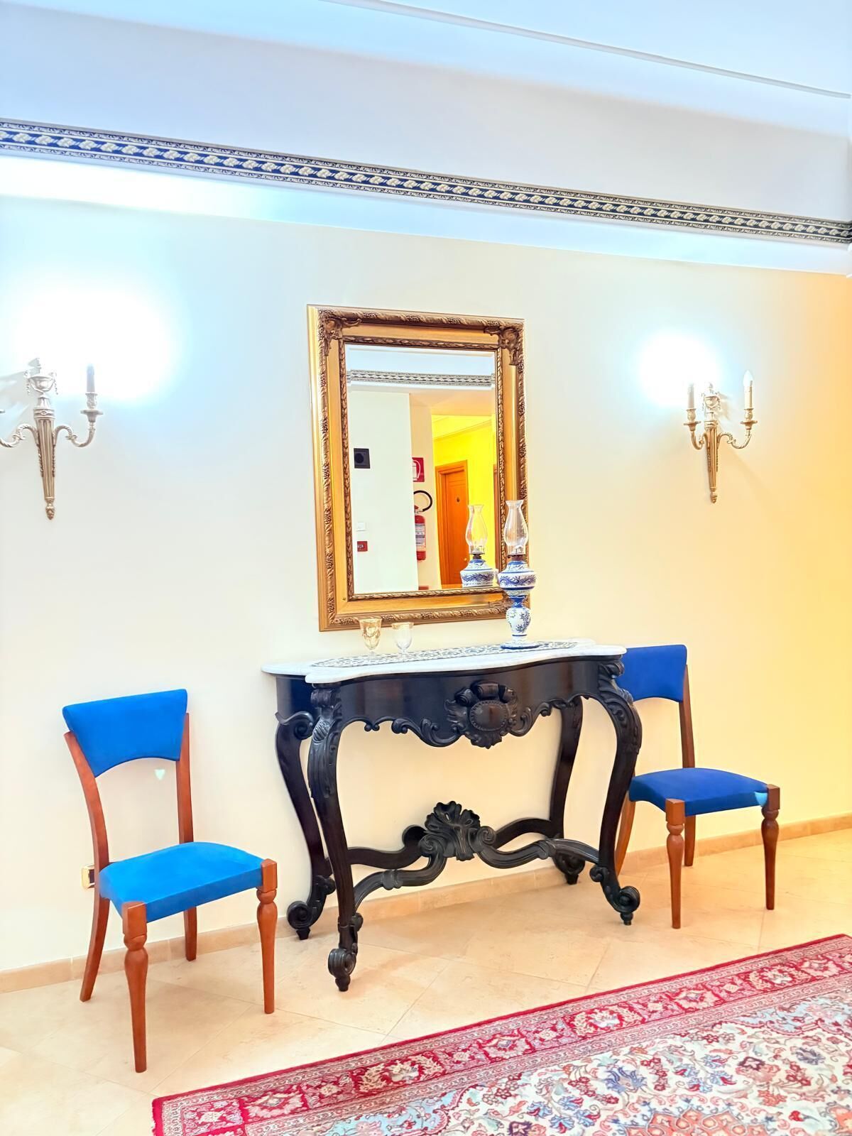 Una habitación con un espejo grande, dos sillas azules, una consola negra y una alfombra estampada.