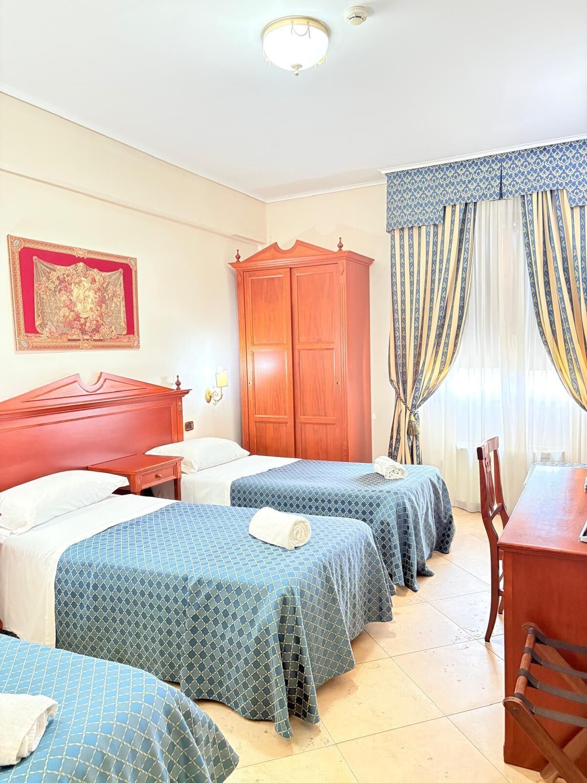 A hotel room with three beds, a wooden wardrobe, a desk, and a chair.