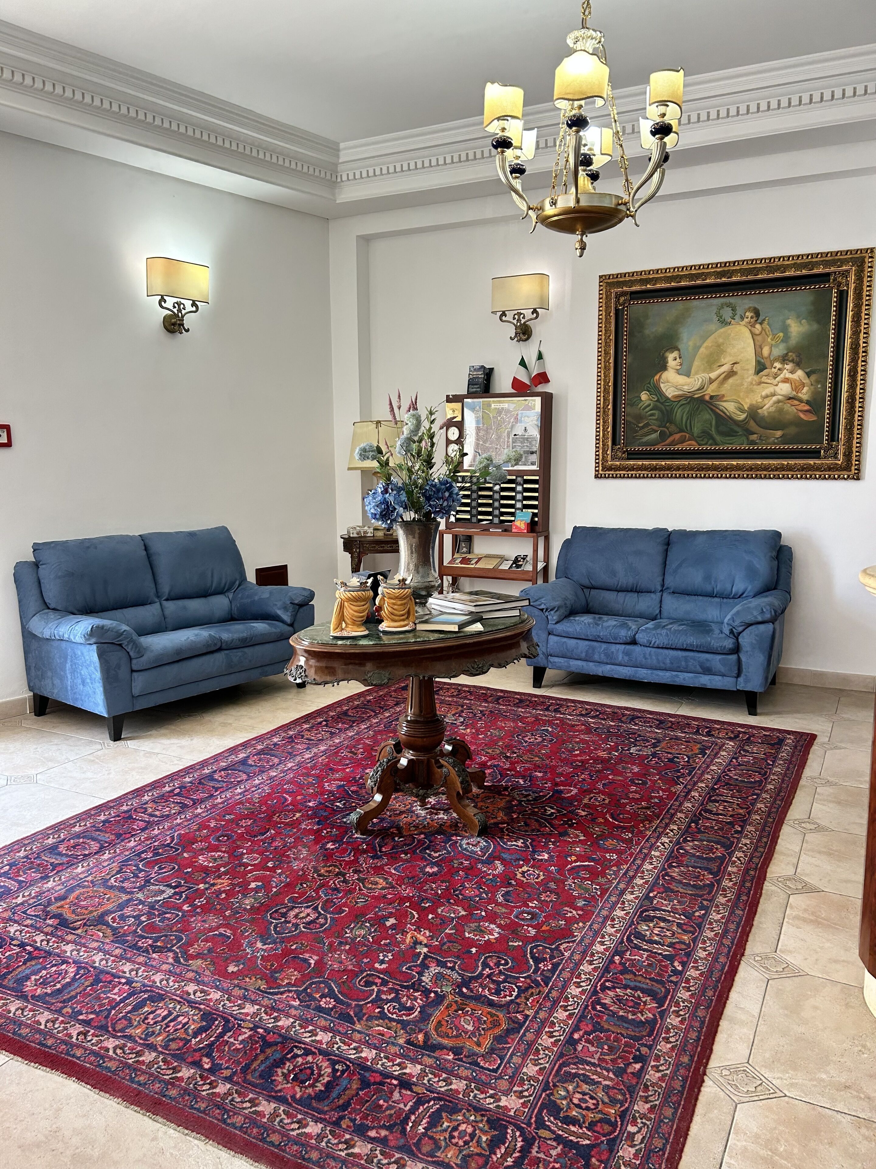 Um quarto com dois sofás azuis, uma mesa redonda de madeira e um tapete vermelho estampado, um candelabro e um quadro na parede.