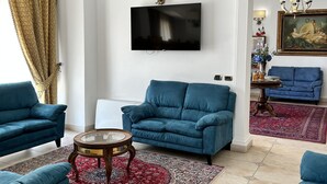 Een ruime woonkamer met een grote flatscreen televisie, blauwe banken, een rood kamerkleed en een kroonluchter.