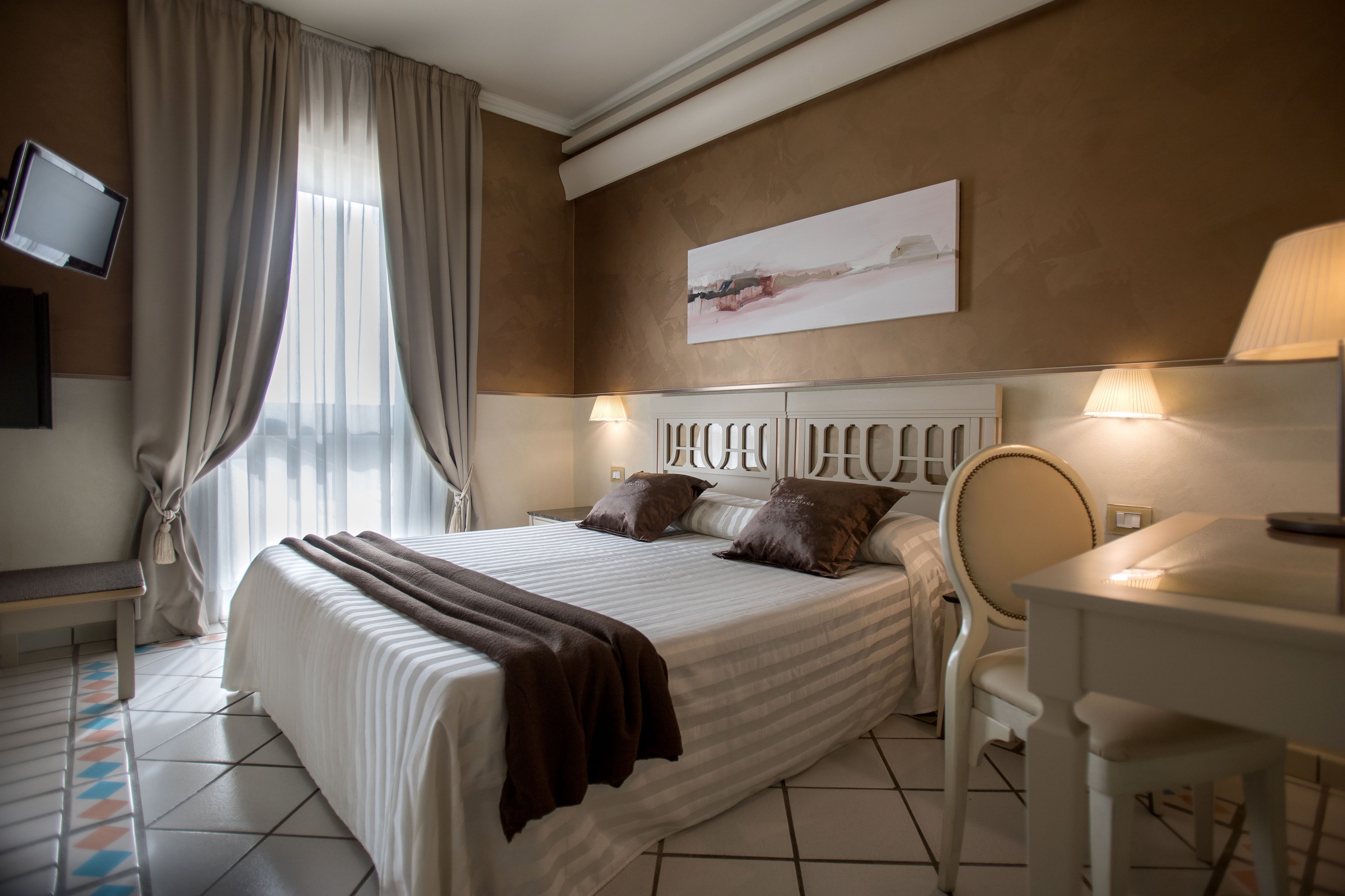 Habitación de hotel con cama, lámparas de noche, escritorio, silla, televisor montado en la pared y un cuadro.