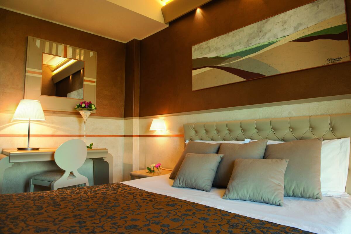 A hotel room with a bed, a desk, a chair, a nightstand, a lamp, and a painting on the wall.