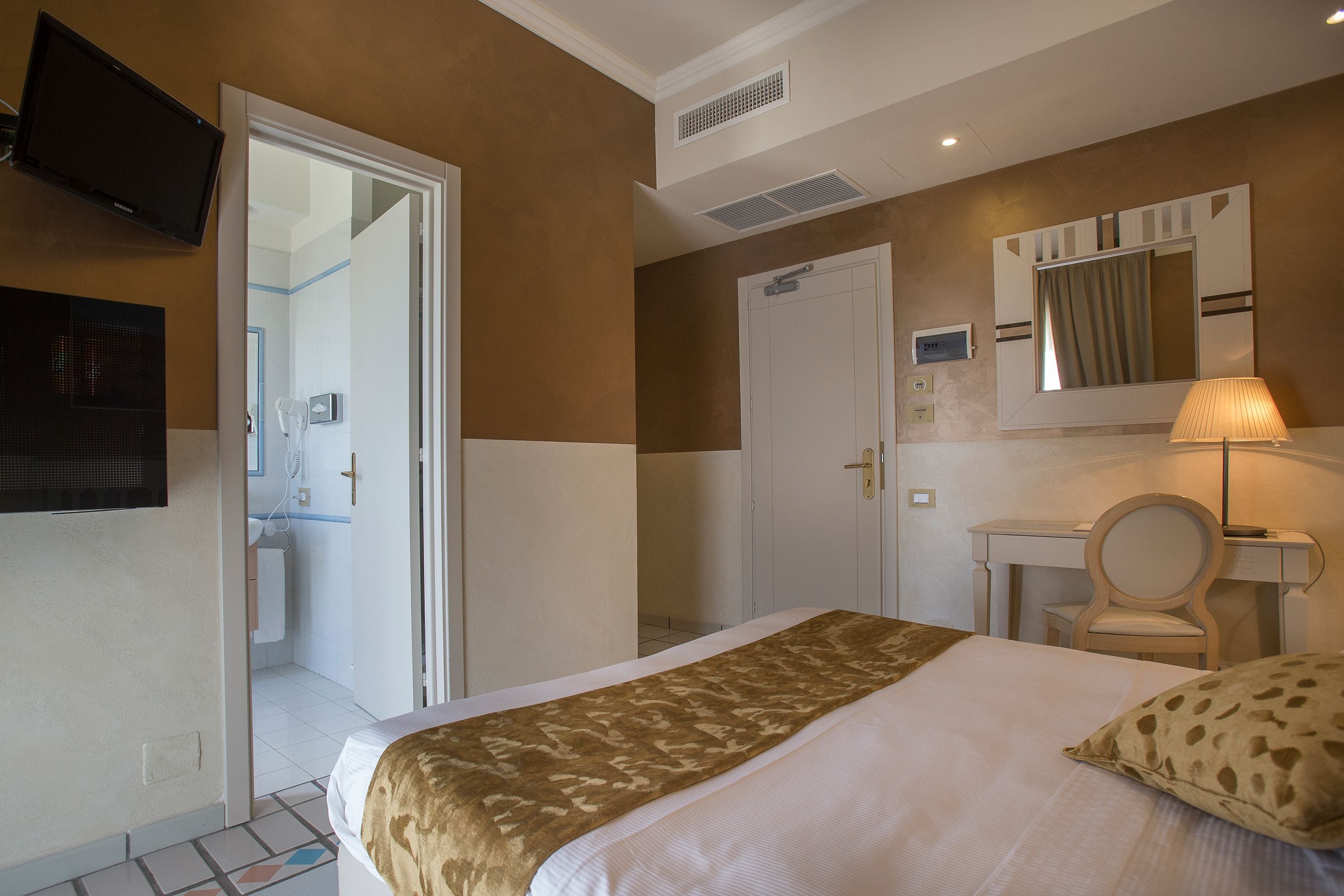 Habitación de hotel con cama, escritorio, silla, televisión y baño visible a través de una puerta abierta.