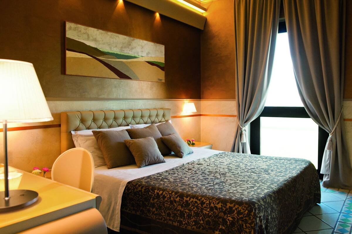 A hotel room with a large bed, a headboard, a bedside table with a lamp, a nightstand, and a window with curtains.