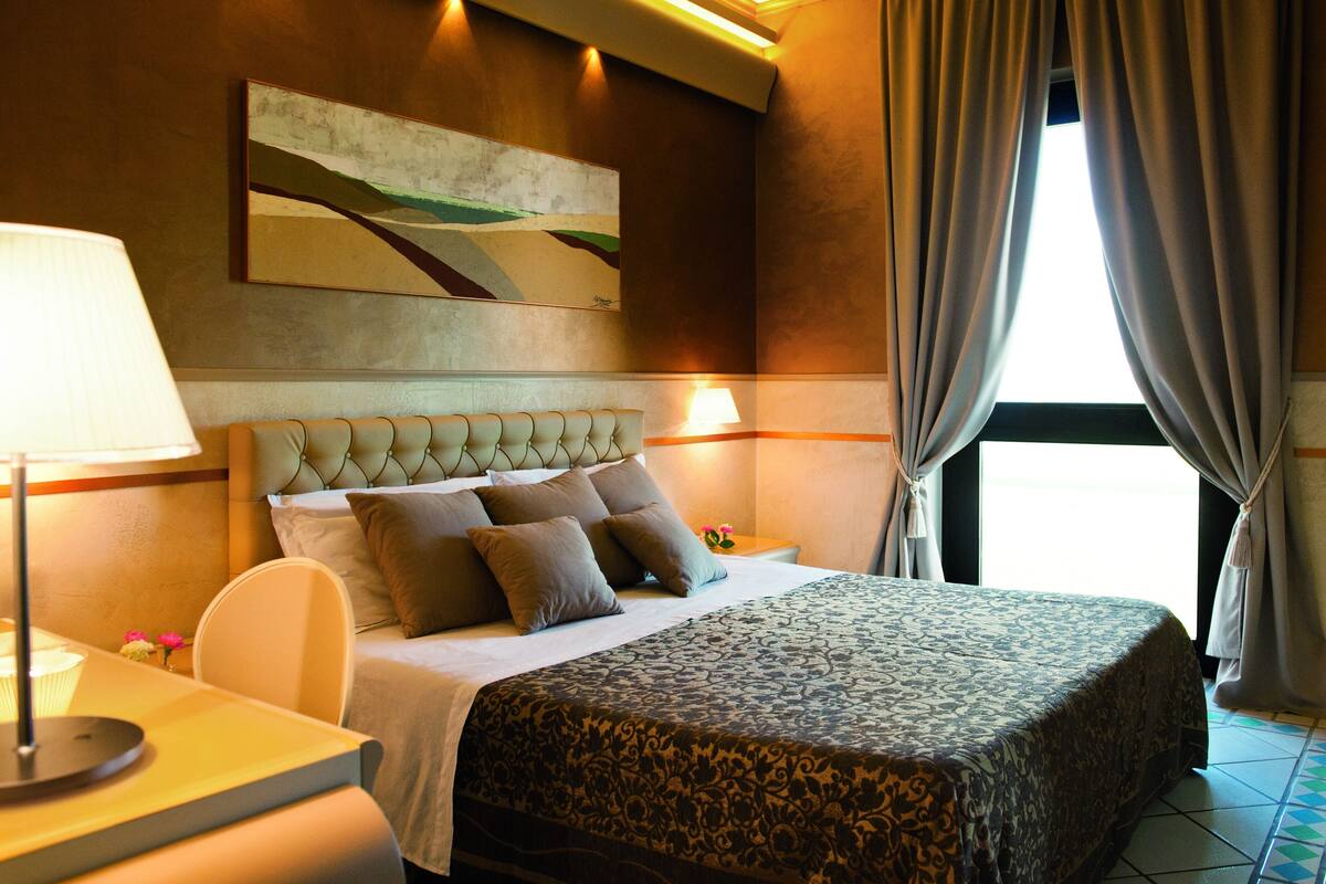 A hotel room with a large bed, a headboard, a bedside table with a lamp, and a painting on the wall.