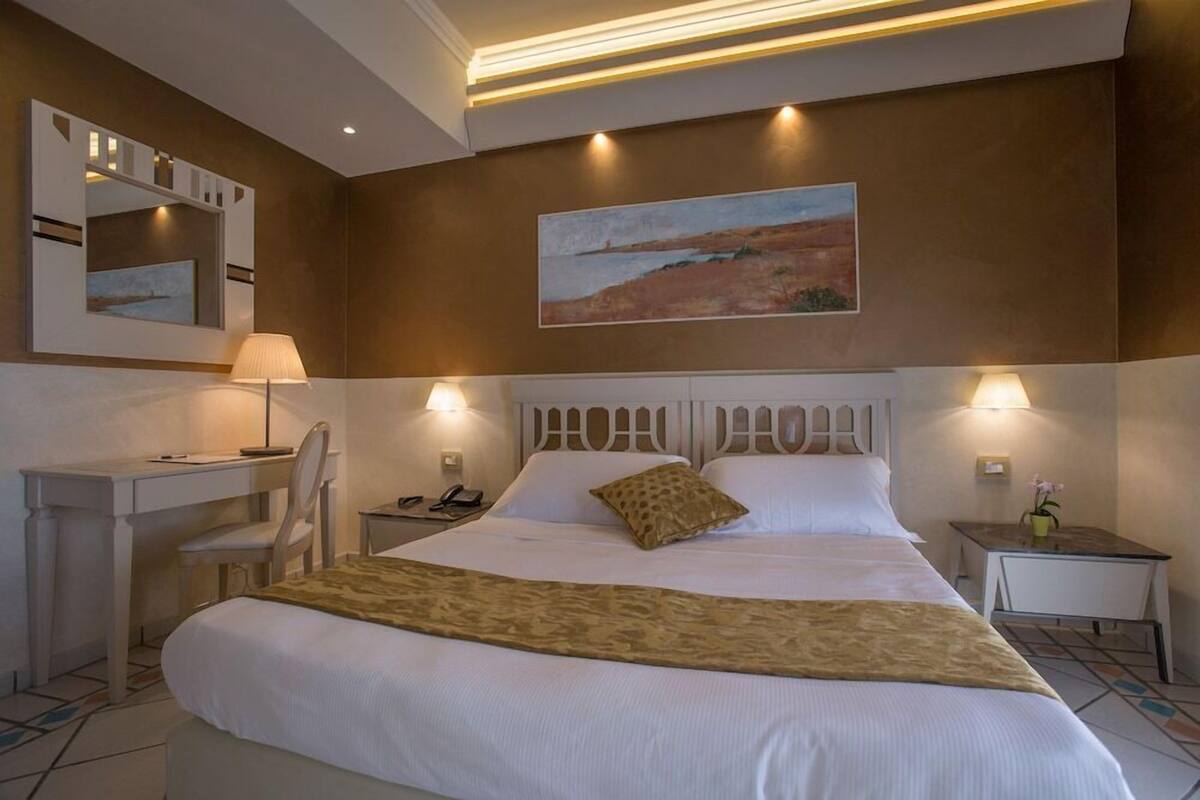 A hotel room with a large bed, a desk, a chair, a nightstand, and a painting on the wall.