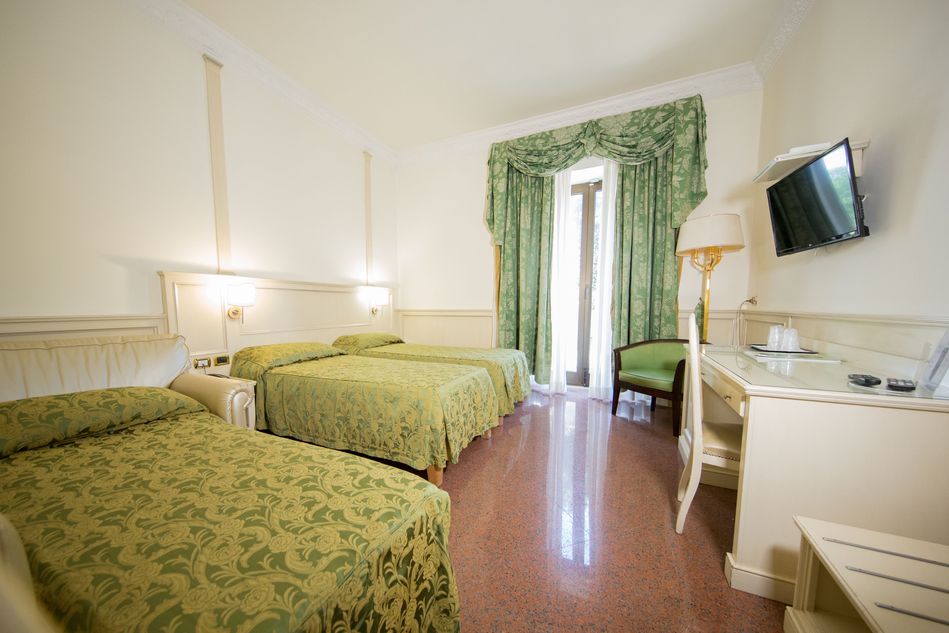 Ein Hotelzimmer mit drei Betten, einem Schreibtisch, einem Stuhl, einem Fernseher und einem Fenster mit Vorhängen.