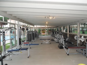 Ein überdachter Fitnessraum mit verschiedenen Trainingsgeräten, Bänken und freiem Blick nach draußen.