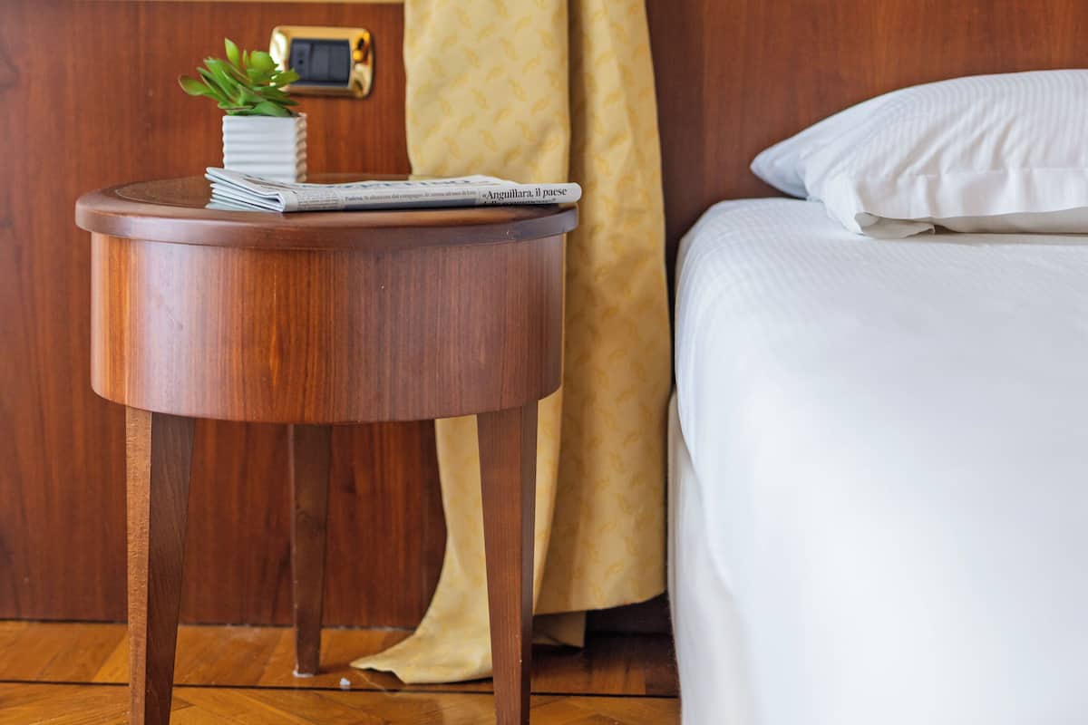 A neatly made bed with white linens, a bedside lamp, a small plant on a wooden side table, and a yellow curtain tied back.