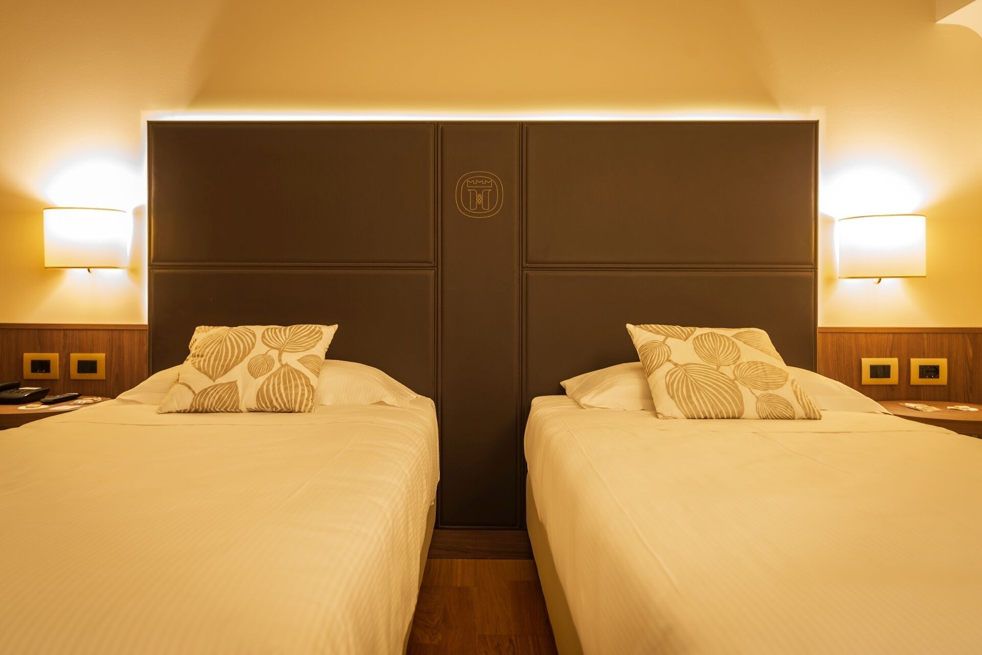 A hotel room with two beds, a dark headboard, bedside lamps, and a small table with a phone and remote controls.