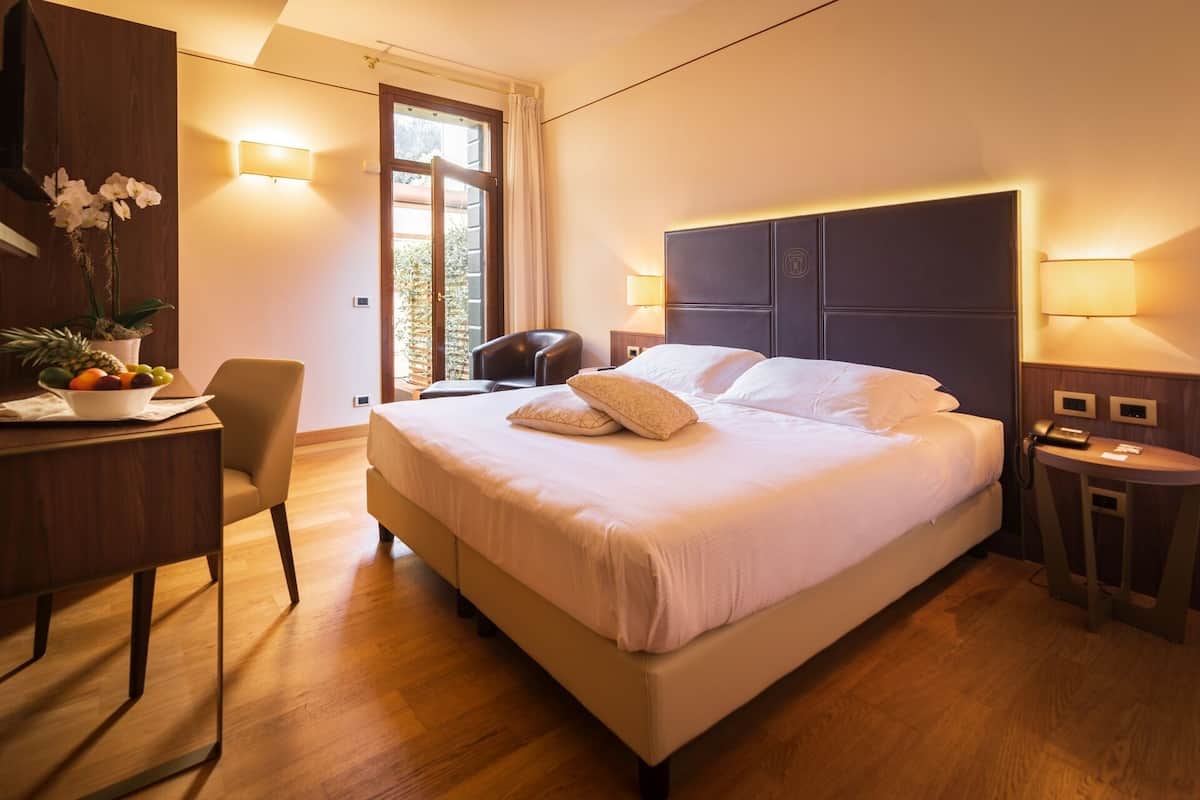 A hotel room with a large bed, a desk with a fruit bowl, a chair, a nightstand, and a window with curtains.