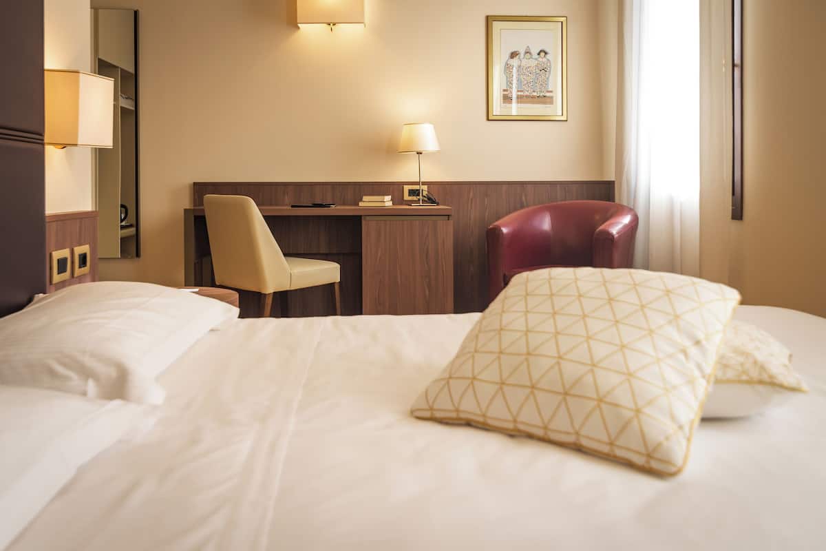A hotel room with a bed, a desk, a chair, a lamp, and a framed picture on the wall.