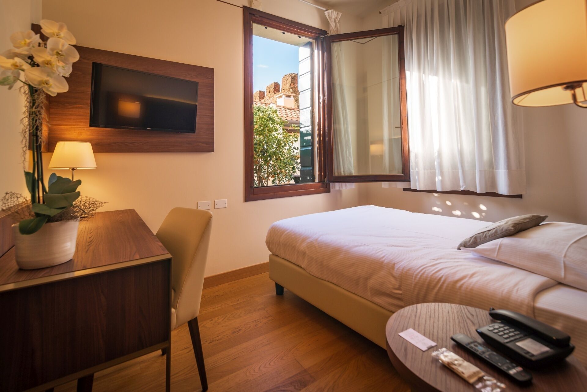 A hotel room with a bed, a desk with a lamp, a TV mounted on the wall, a window with curtains, and a view of buildings outside.