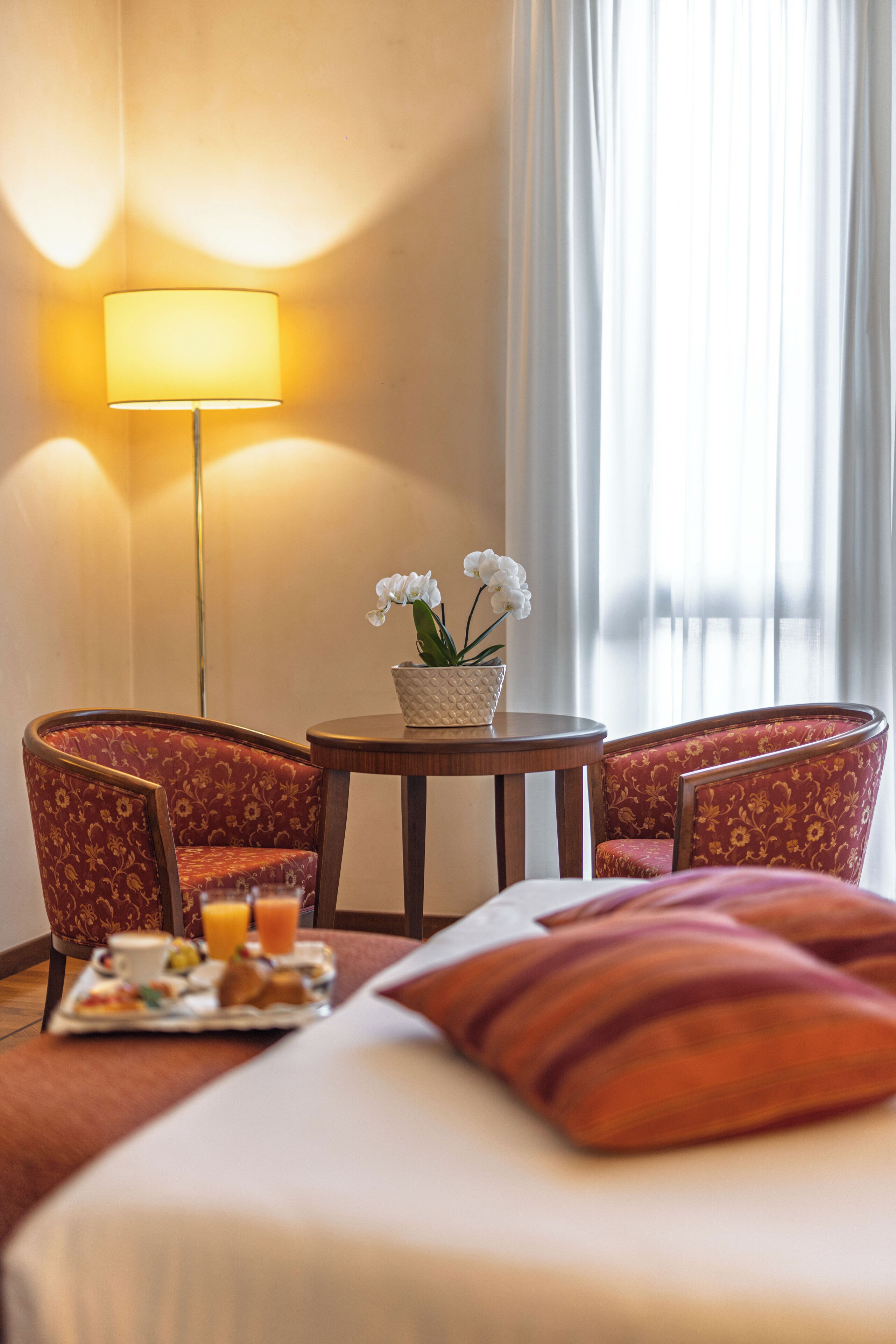 A hotel room with a bed, two armchairs, a small table with a flower vase, and a tray with breakfast items.