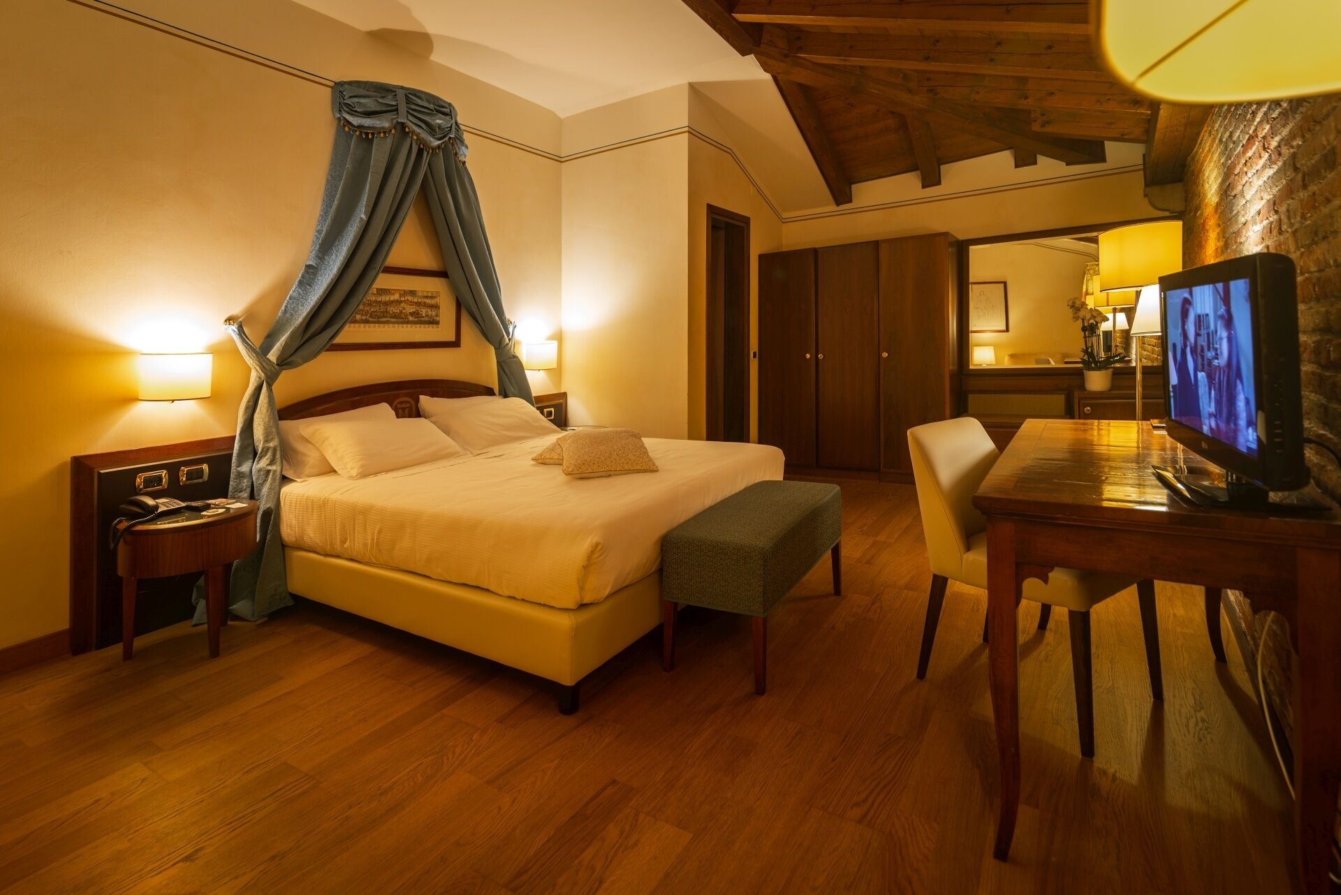A hotel room with a bed, bedside table, lamp, TV, and a wooden desk.