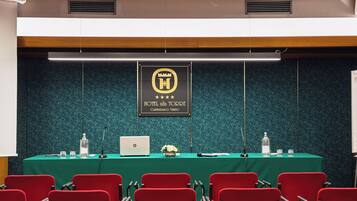 Una sala de conferencias con una mesa verde, sillas rojas, una computadora portátil y un micrófono. Se ve un cartel con el nombre del hotel.