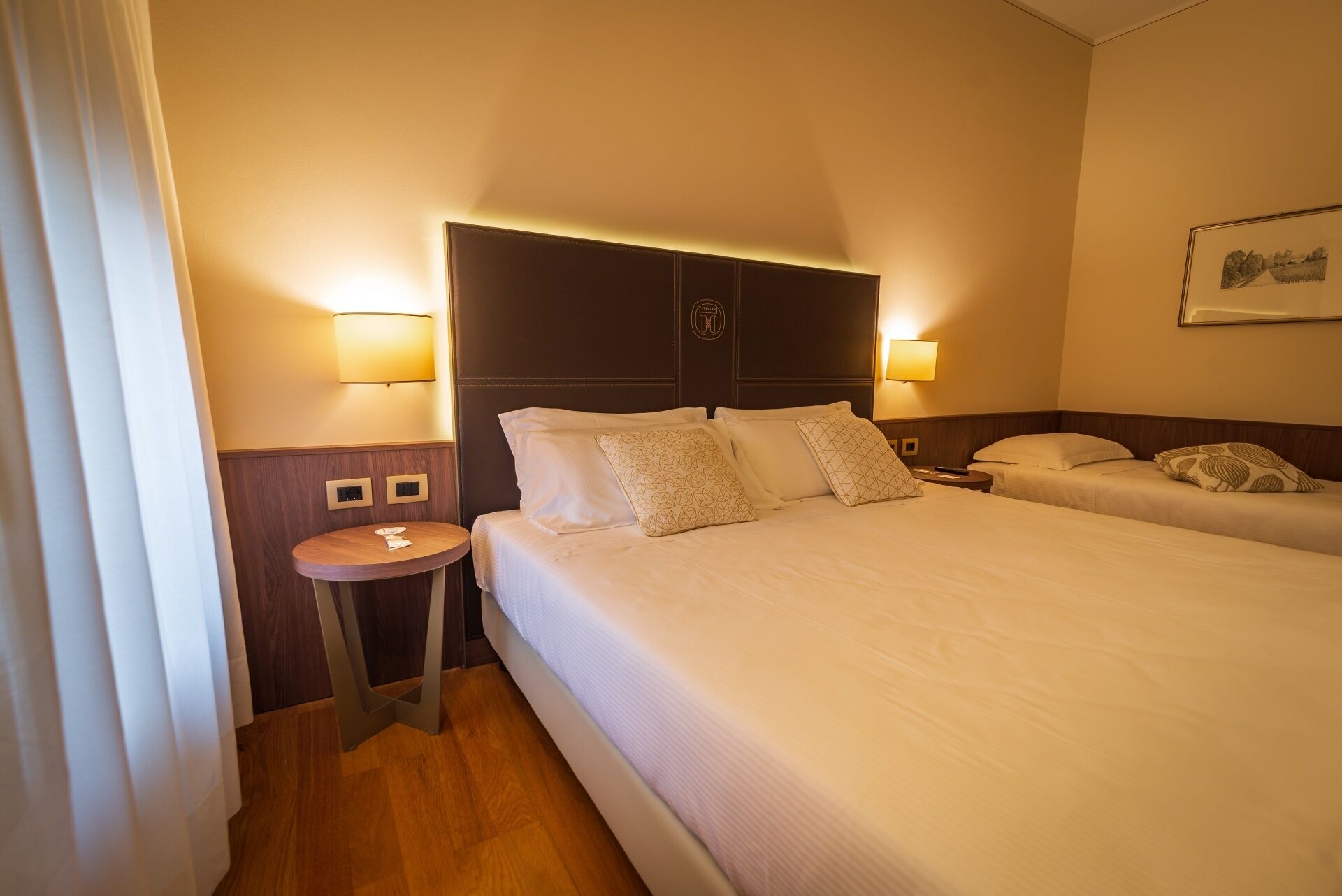 A hotel room with two beds, a round table, and wall-mounted lamps.