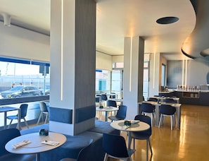 Un restaurante moderno con mesas redondas y sillas azules, pilares grandes y vista a la calle.