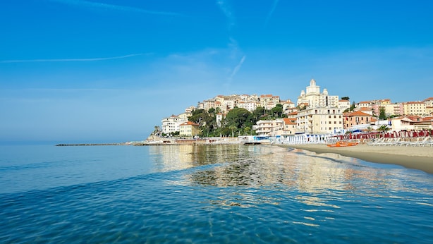 Un paysage urbain côtier avec des bâtiments le long du front de mer, une eau d’un bleu limpide et une église imposante.