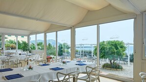 Un restaurant avec des tables et des chaises blanches, de grandes fenêtres donnant sur la mer et la verdure.