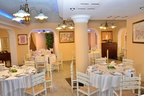 A banquet hall with round tables set for dining, white chairs, and chandeliers. - Cala Ginepro Hotel Resort (Orosei)